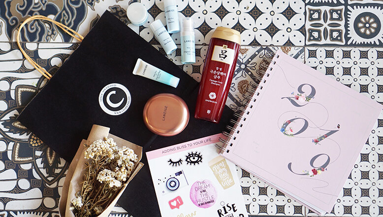 Goodie bag contents A small flower bouquet, stickers, a scrapbook, a foundation cushion, a bottle of shampoo, a black paper bag, and other sorts of skincare products are all laid down on mosaic tiles.