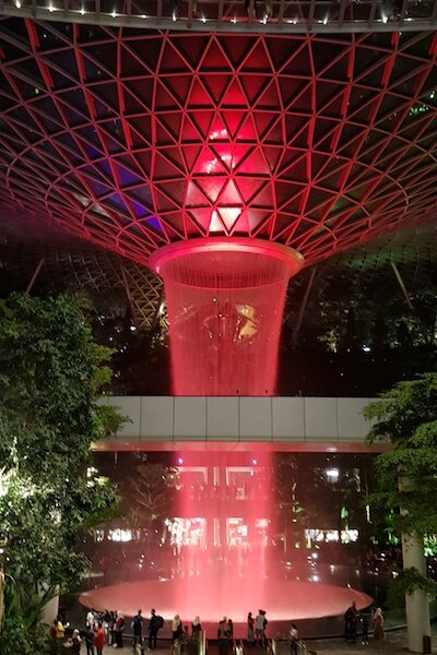 A tall indoor waterfall illuminated by red light and surrounded by greenery