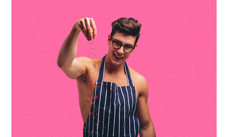 The Topless Baker wearing a navy striped apron sprinkling multi-coloured sprikles.