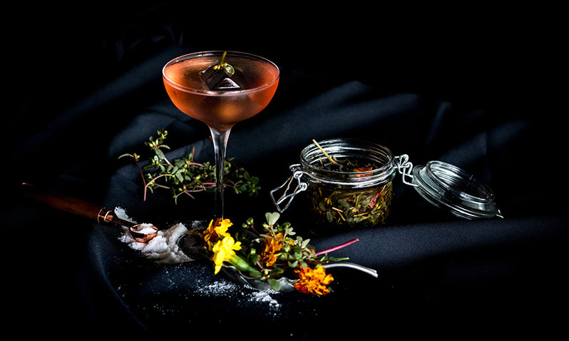 A glass of a cocktail surrounded by flowers, salt, and other ingredients