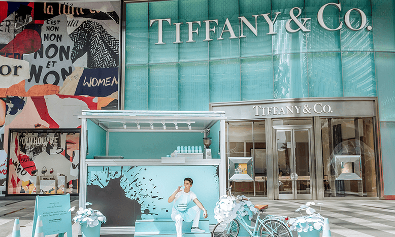 A photo of a man sipping a drink in a coffee cart completely coloured in Tiffany blue right in front of a Tiffany & Co store.